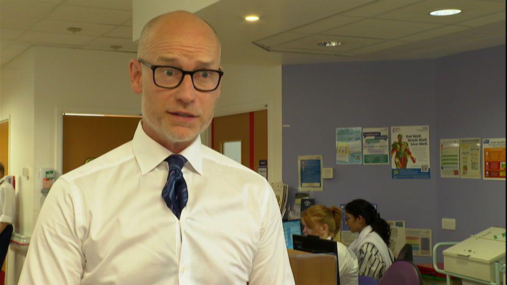 A bald man with black glasses and a white shirt, with a blue tie tucked into the space between the buttons, stands on a hospital ward. Two women sit at a desk looking at computers behind him.