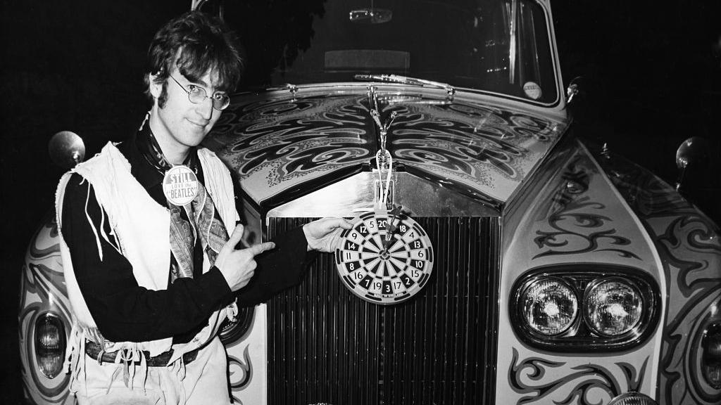 A black and white photograph of John Lennon smiling with his psychedelic Rolls-Royce