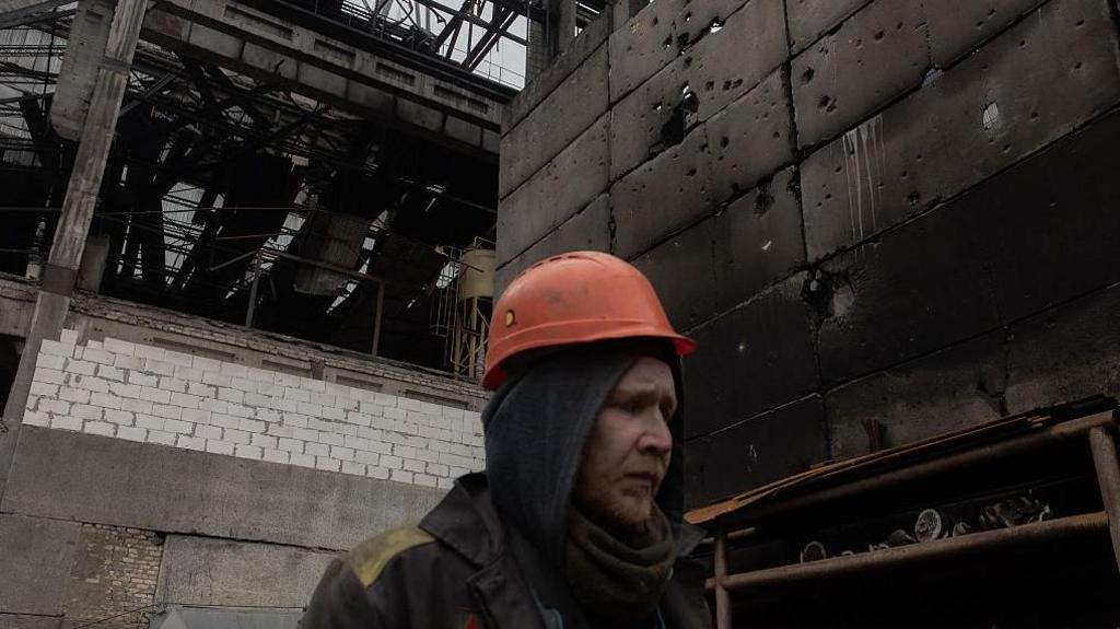 A workman in an orange safety helmet walks through a large charred industrial plant.