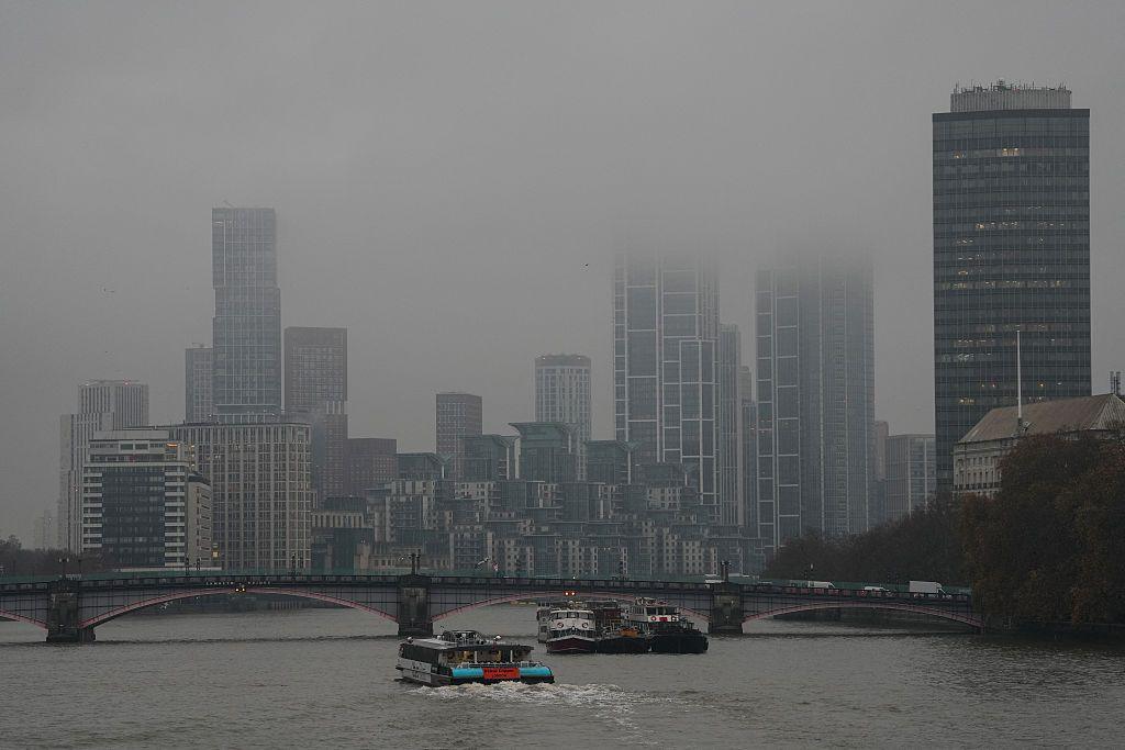 A catamaran makes it way down the Thames towards high-rise buildings partly obscured by clouds in London