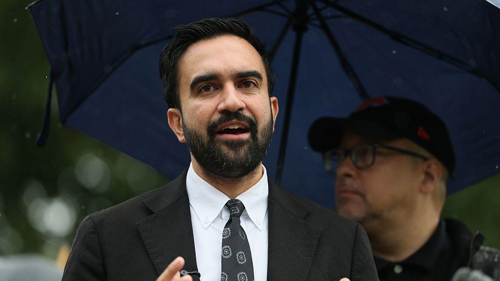 Zohran Mamdani in a black suit and black tie under a large blue umbrella being held by a police officer.