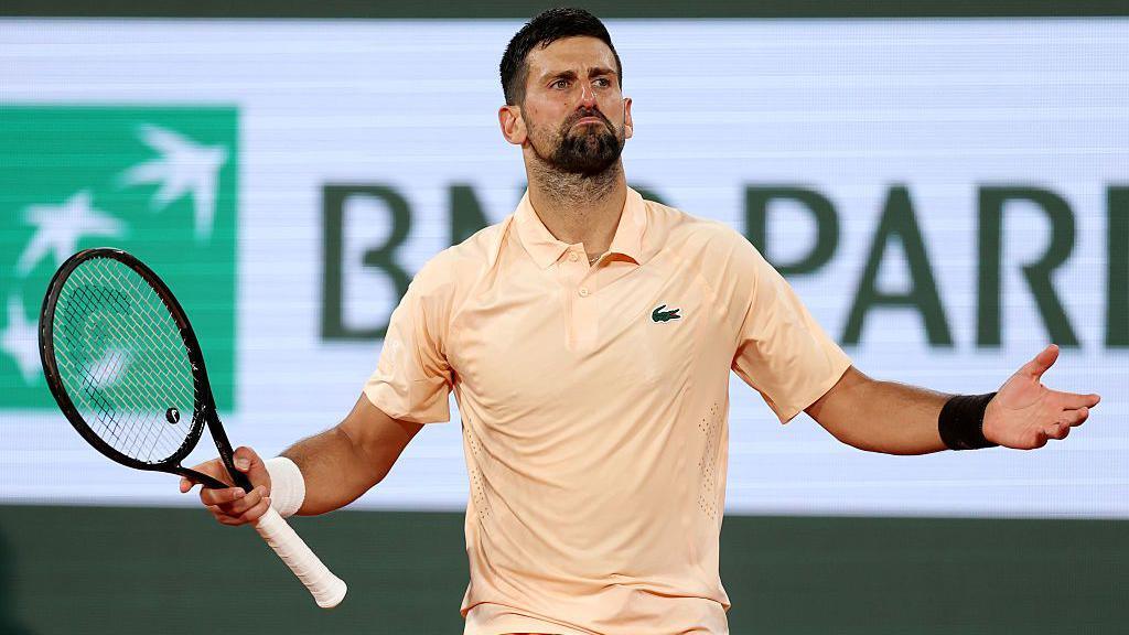 Novak Djokovic, wearing a peach polo shirt, holds his arms wide with his racquet in his right hand
