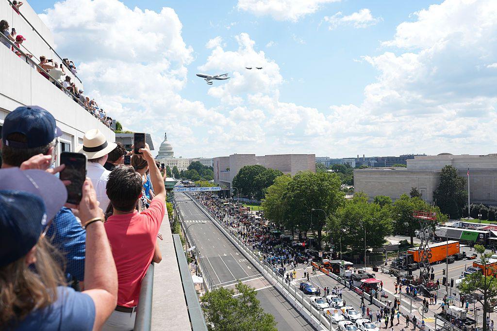 People take photos on a rooftop as Air Force one takes part in a flyover with the Capitol building in the background and the racetrack below