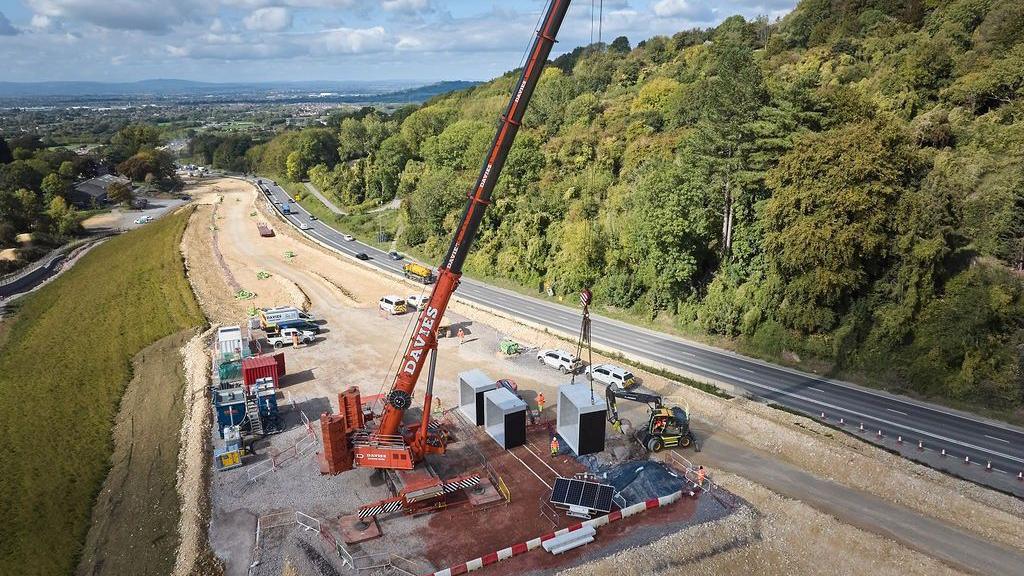 A bird's-eye view of the bat tunnel being installed under the road. A large red crane is visible on the building site which is next to a road surrounded by fields and trees.