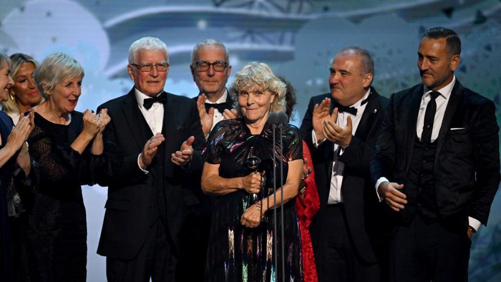 Jo Hamilton, a woman with grey hair in a shiny black dress, holds a trophy whilst being clapped and cheered by actors and producers around her on the stage