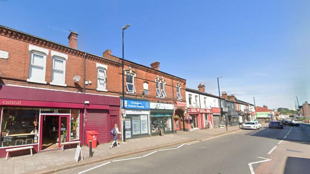 A high street on a sunny day. There are two-storey buildings on the left side of the street with colourful shops and cafes on the ground floor level. There is one person walking down the street.