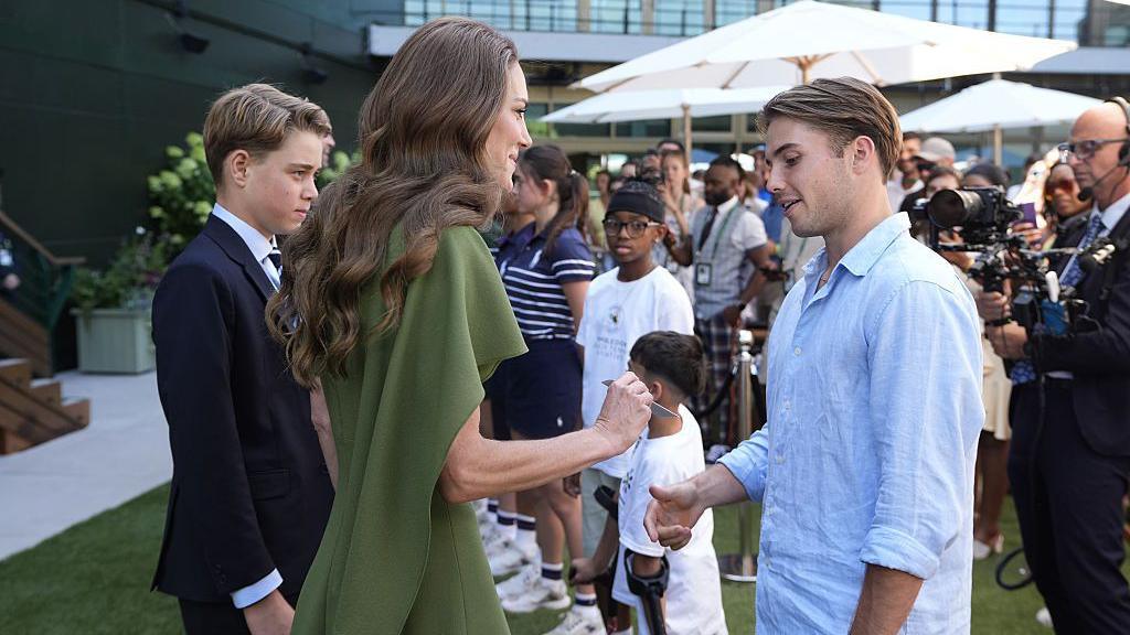 Princess of Wales is seen wearing a green dress and handing an envelope containing a birthday card to Arthur Fery, seen wearing a blue shirt. Prince George stands next to his mother wearing a black suit. 