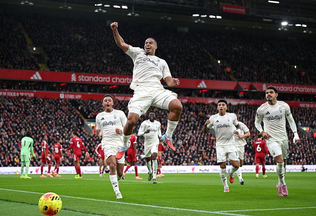 A Nottingham Forest player leaps in celebration after scoring, with team-mates rushing toward him as the crowd fills the stadium and Liverpool players look on in the background.