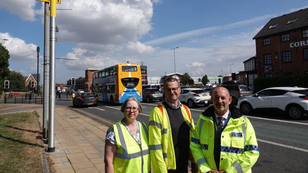 A woman and two men stood on a path next to a road with cars and buses on. They are all wearing hi-vis jackets.