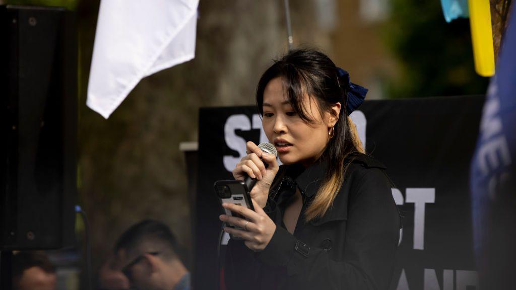 Carmen Lau wearing a black coat and blue hair bow reading a speech from her phone at a protest outside Downing Street in 2022.