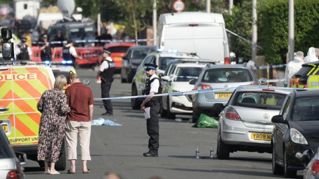 Members of the public look on from in front of blue and white police tape as police officers stand in a road filled with police vans and cars, with debris strewn across it.