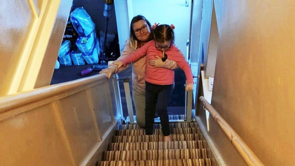 Picture taken from the top of a house's stairs, looking down to the bottom, where Claire is pictured holding her daughter up with her arm across her chest, as she tries to climb. Claire is wearing a grey tracksuit with glasses, while Emilia has black leggings and a pink long-sleeved t-shirt.