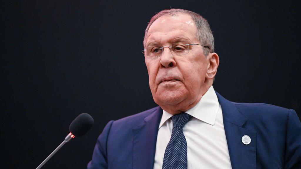 Sergei Lavrov sits in front of a small black microphone. He is wearing a blue jacket, a white shirt and a spotted tie.