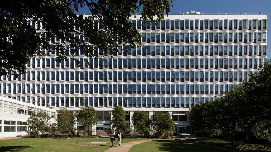 A large mid-rise building of white concrete and blue windows.The building facade is seen from a grass courtyard that also features multiple short trees.