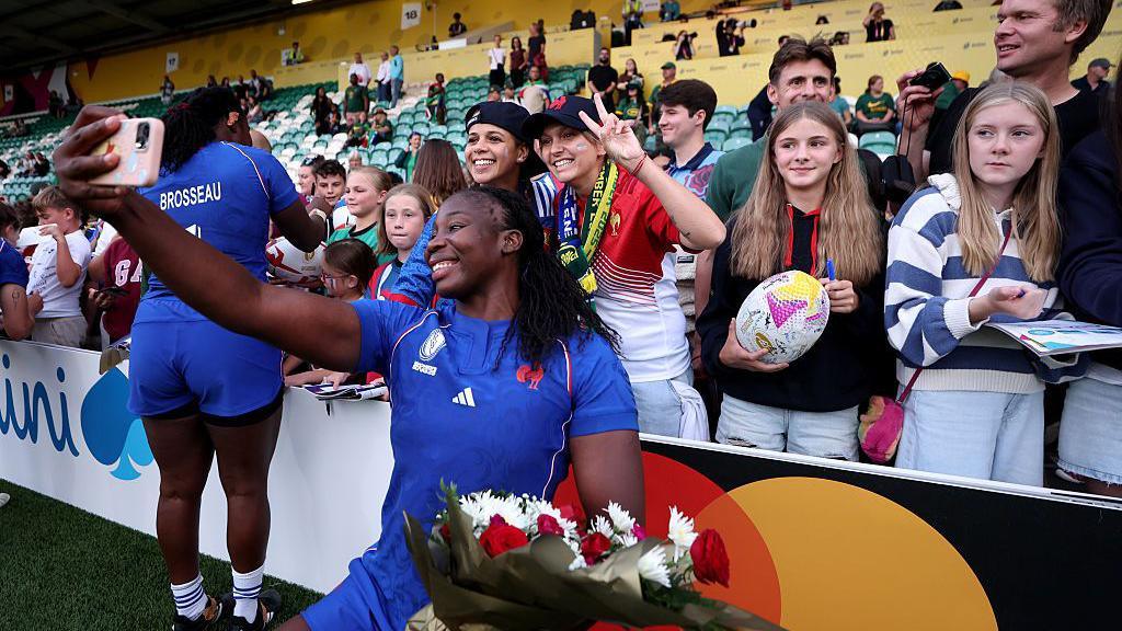 A French rugby player poses for a selfie in front of fans. She is holding a bunch of flowers. The player and the fans are smiling.