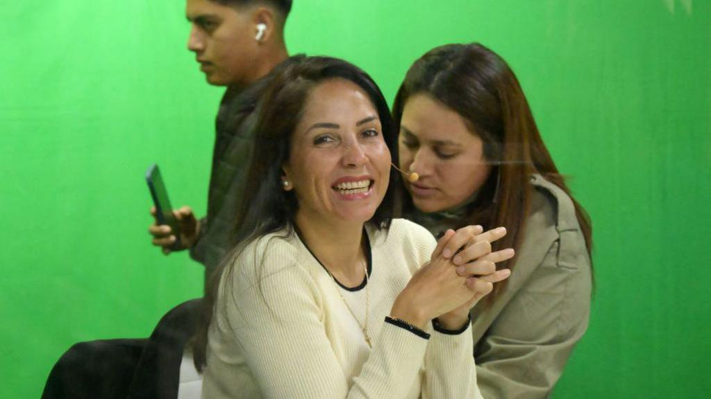 Luisa González sits down, smiling with her hands clasped together as two people stand behind her