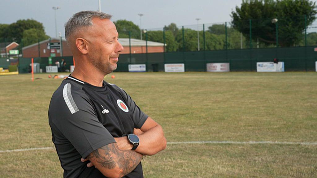 Andy Crowther stands with his arms folded, looking away from the camera and watching something in the distance. He is wearing a black T-shirt with Droitwich Spa branding and short grey hair and stubble.