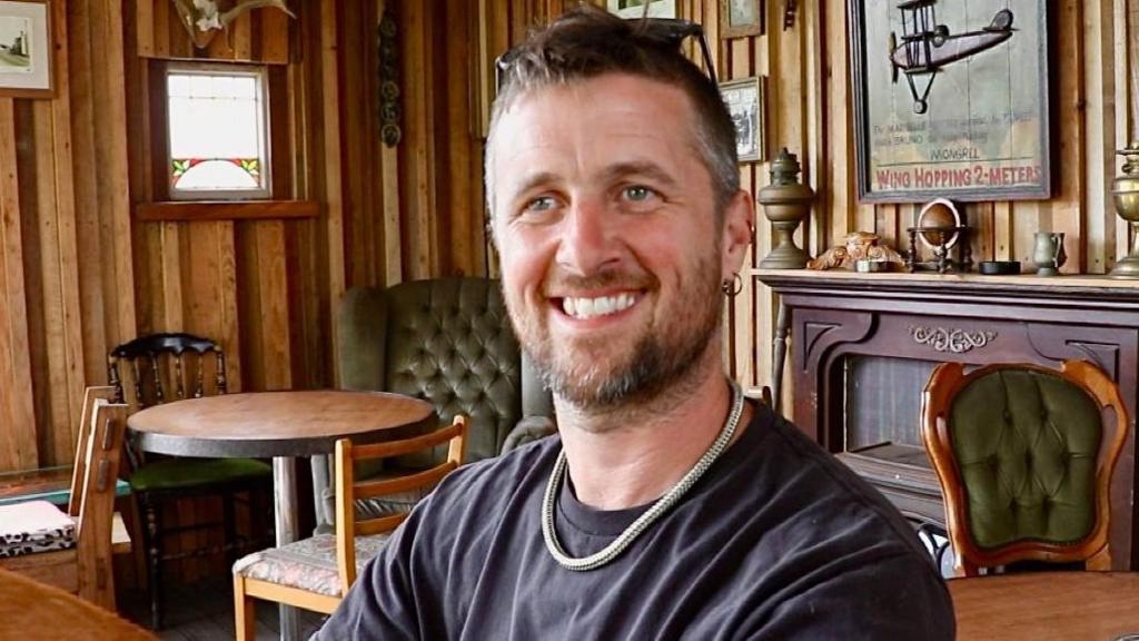 Owain Powell sitting and smiling in his pub. There is a lot of wooden furniture and the walls are lined with wooden slats