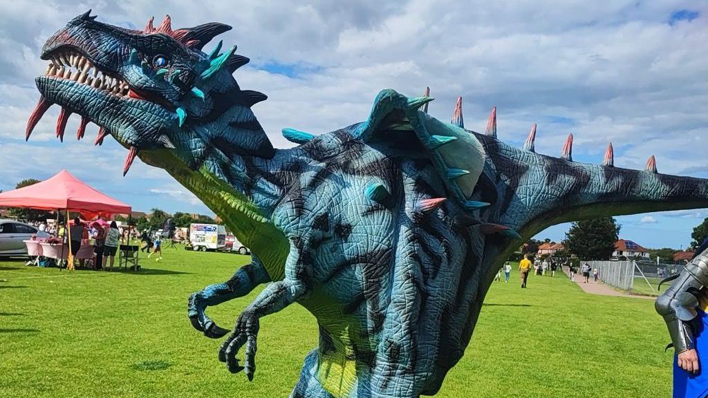 A large dragon puppet with red spikes on its back and sharp teeth walking on a field