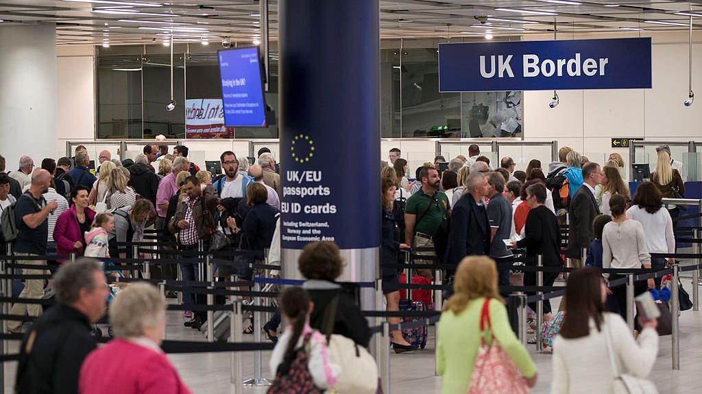 The UK border control area at Gatwick Airport. The white-walled hall is busy with people who are queuing in a long, winding line. Some are holding suitcases and passports. There is a large blue column in the middle of the room which says: "UK/EU passports" and a large sign hangs from the ceiling which reads "UK Border".