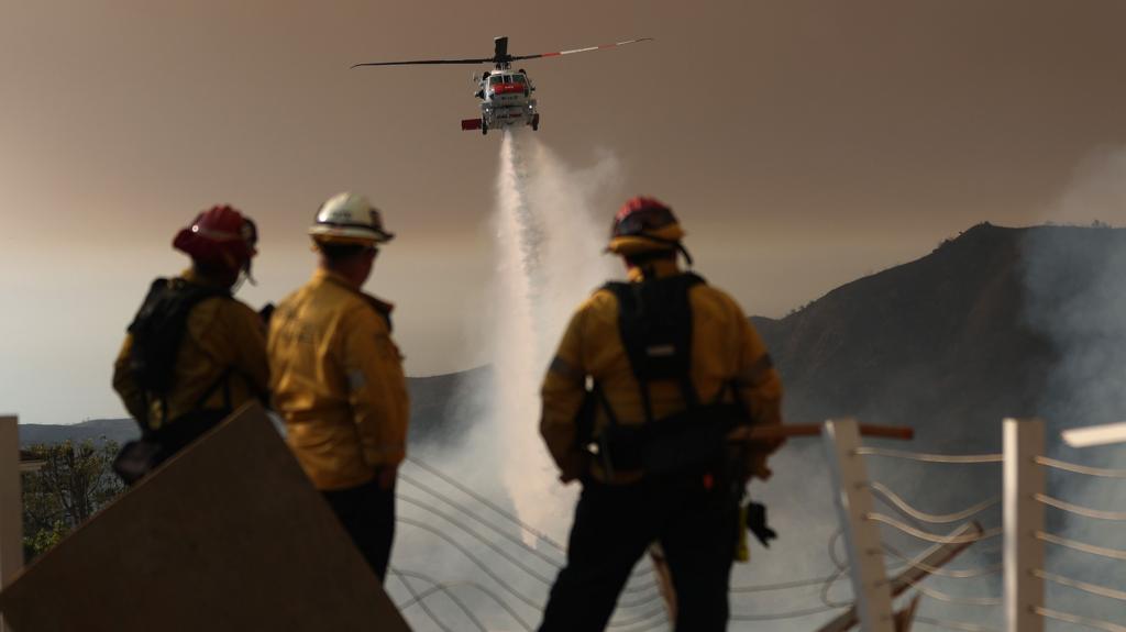 Three firefighters in protective gear watch a firefighting helicopter dump a payload of water on a wildfire