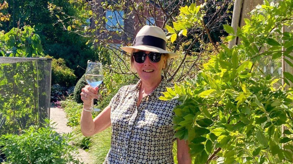 Sally Hall holds up a wineglass in a garden on a sunny day wearing black shades wearing a straw hat. 