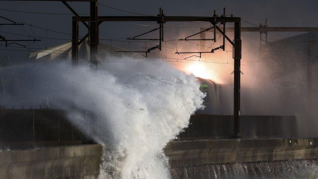 A train is on a track right by a sea wall. Water is being pushed upwards and over train carriages. The water has struck the overhead wires causing them to spark dramatically.                                                                                                                                                                                                                                                                                                                                                                                                                                           