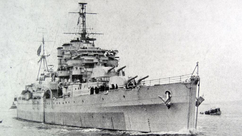 Old Black and while image of large metal navy ship in the sea, with large guns and masts.
