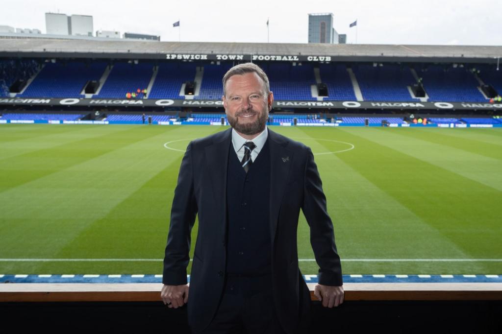 Mark Ashton leaning against a barrier in a stand at Ipswich Town, with the pitch behind him. He is wearing a dark suit, pale shirt and tie.