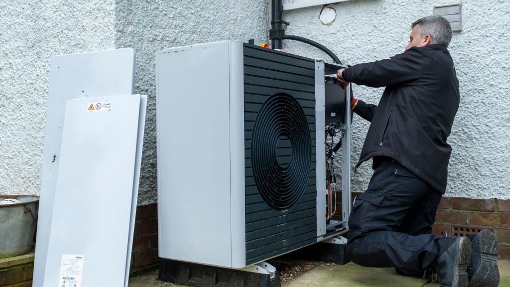 An engineer is installing a heat pump on the side of a property. He is wearing a black jacket, trousers and boots. The building behind him is white and the large heat pump unit is visible in the foreground.