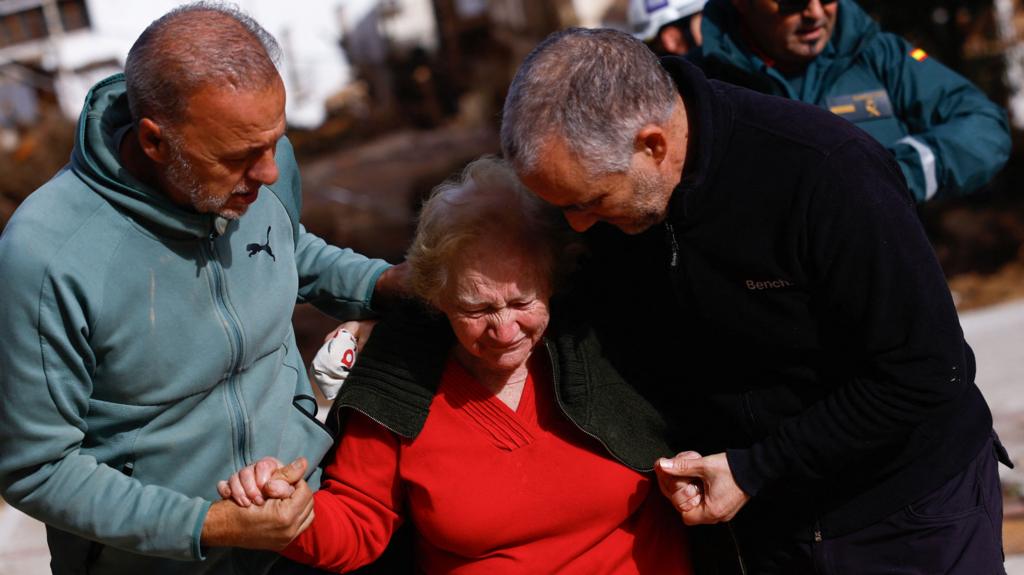 An elderly woman cries as two men, with their arms around her, hold her hands and lead her away