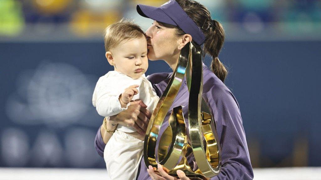 Switerland tennis star Belind Bencic, wearing a purple tracksuit top and visor, holds her baby Bella in one hand and a golden trophy in the other, planting a kiss on the side of Bella's head.