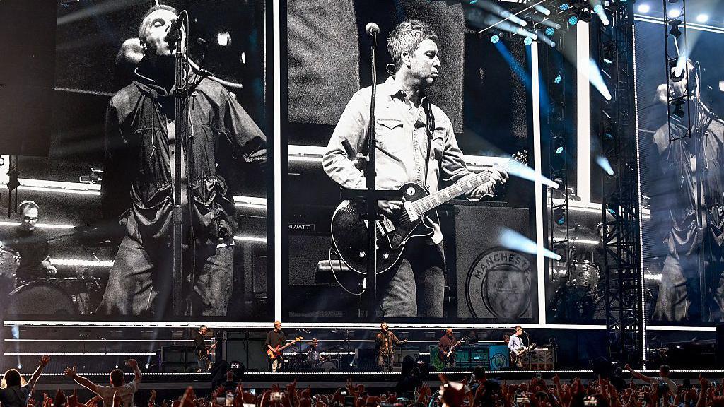 Oasis band stand on stage at Principality stadium Cardiff. Close up shots of the brothers can be seen on large screens behind them. Hands can be seen raised in the air in the crowd. 