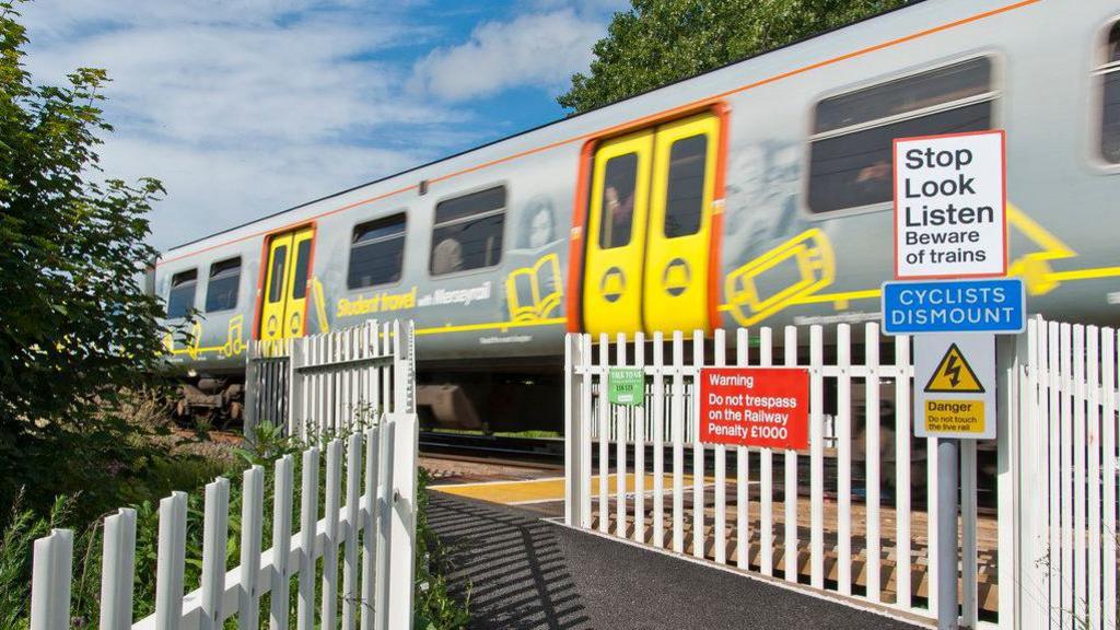 Train passes fast at level crossing with white gate open. Sign says Stop, Look, Listen. Beware of trains.  Cyclists dismount. Another red sign says warning do not trespass on the railway. Penalty £1000