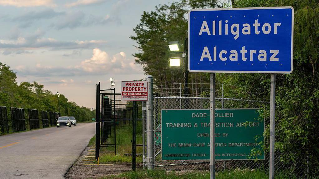 A blue sign saying 'Alligator Alcatraz' at a road entrance followed by a long road, lined by metal security fencing.