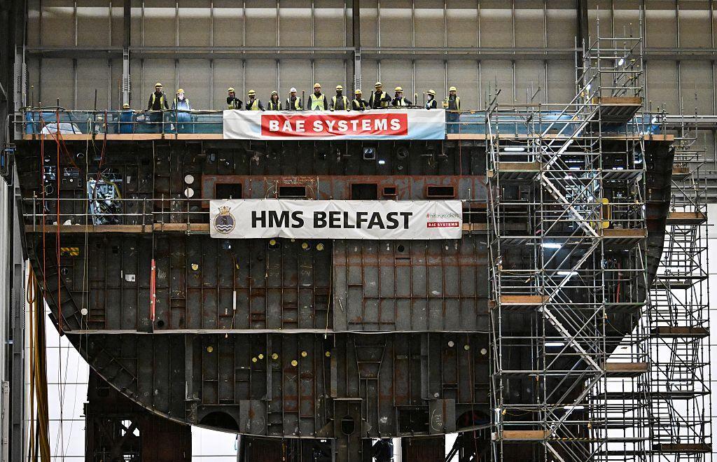 A large warship, under construction, with workers on the deck and scaffolding at the side. The ship is in profile so the interior compartments are visible