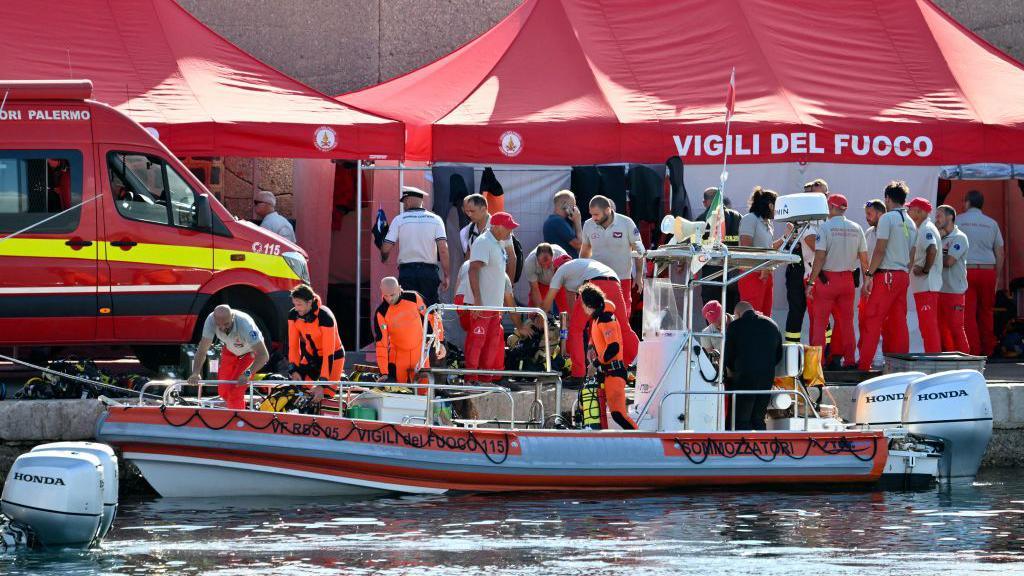 Red tents, a van and a boat belonging to the Italian fire service at the waterfront with a concrete wall behind them. Lots of firefighters, divers and other uniformed officials are milling around on land and standing in the boat.