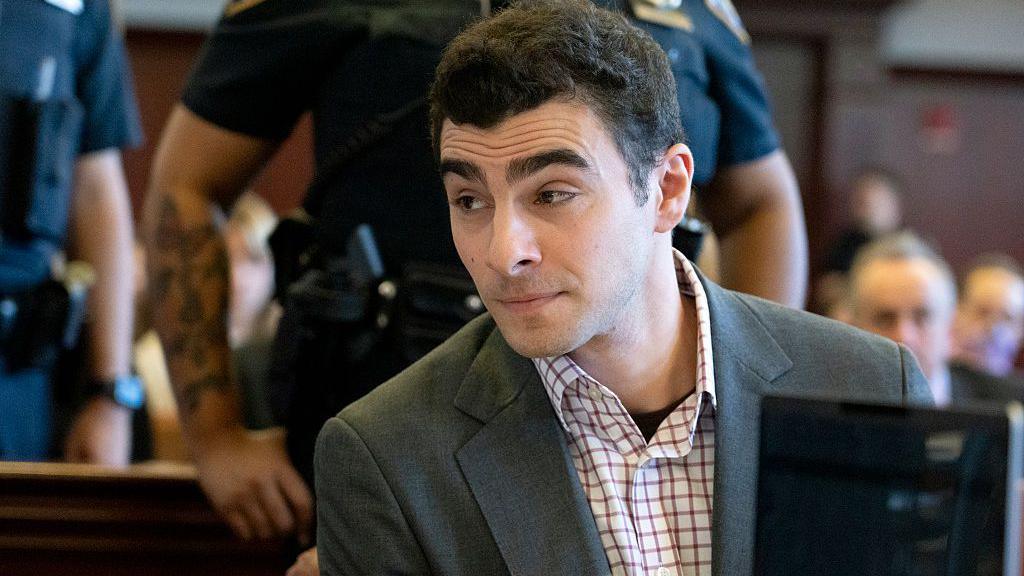 Mangione, wearing a blazer and button-down, looks off to the side in courtroom with police officer standing behind him