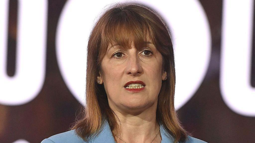 Chancellor Rachel Reeves, wearing a blue jacket, stands in front of a bright white sign, with the o in the sign surrounding her head.