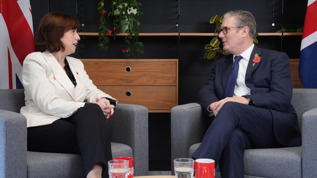 Lucy Powell (L) in white blazer, black top and trousers sits on a grey armchair while speaking with Keir Starmer (R) in a dark blue suit sitting on a matching chair