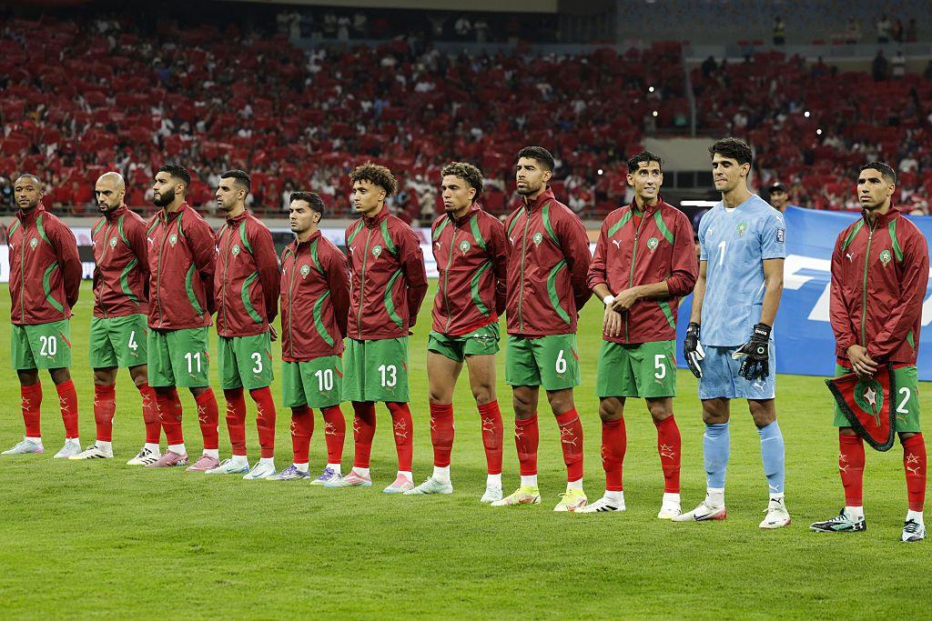 Morocco's men’s national football team lined up, during the national anthem, on the pitch before a match. There are 11 players standing shoulder to shoulder in a straight line. Most are wearing Morocco’s traditional home colours: red jackets with green detail, green shorts and red socks. The goalkeeper stands out in a light blue kit with gloves on, positioned slightly apart but still aligned with the team. A packed stadium forms the background, with large sections of red-clad supporters filling the stands, reinforcing a home or strongly supported setting