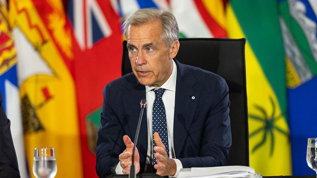Mark Carney, in a blue suit and polka dot blue tie, is seated in a char and speaks into a microphone. Behind him are a number of flags hanging in multiple bright colours, mainly yellow, red, blue and green.