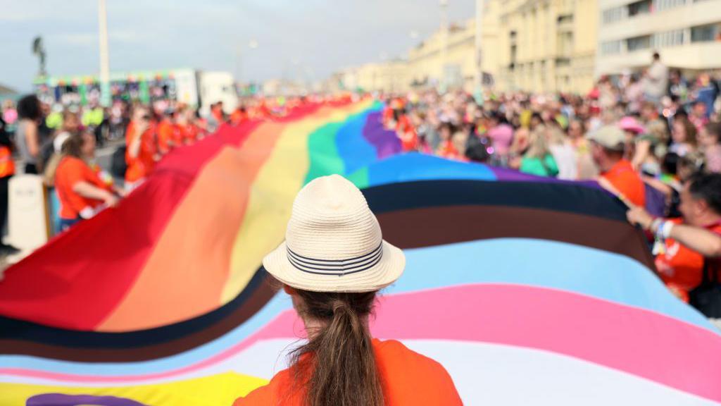 A woman in a hat stands staring at a long rainbow flag draped along a street.