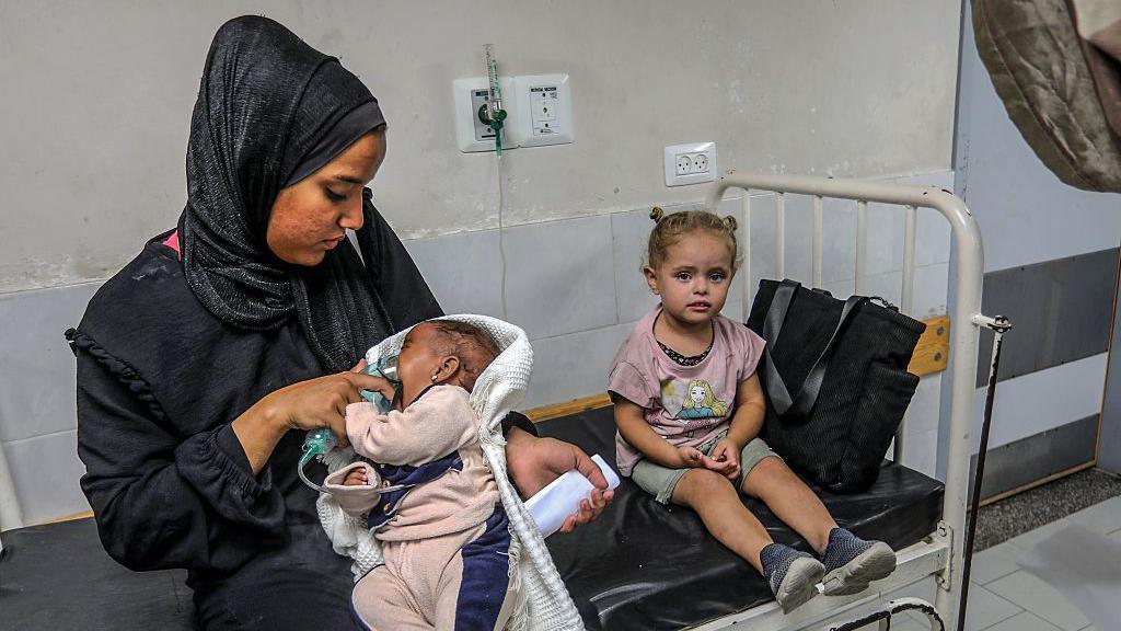 A Palestinian woman nurses a child in her lap on a hospital bed.