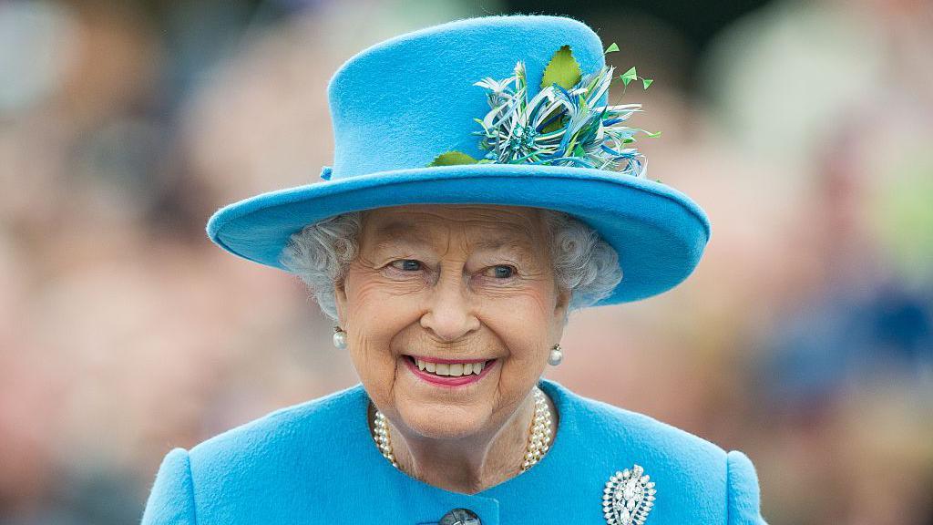 Queen Elizabeth II pictured in 2021 in a blue jacket and blue hat - she smiles and wears pink lipstick