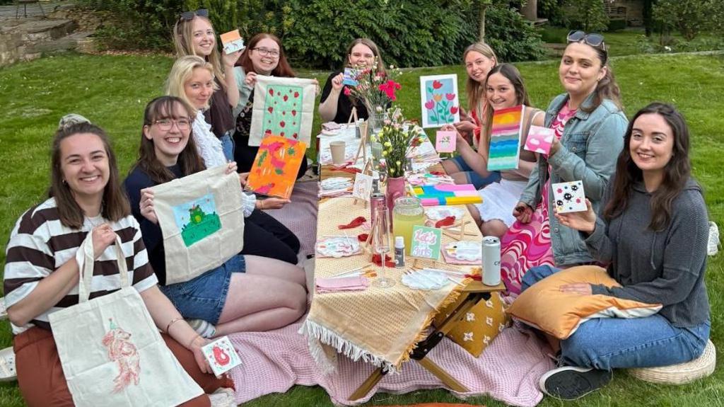 A group of young women sitting around a low lying table holding up colourful embordered bags they have made. The table is set with lots of colourful table cloths and flowers. They are all smiling at the camera. 