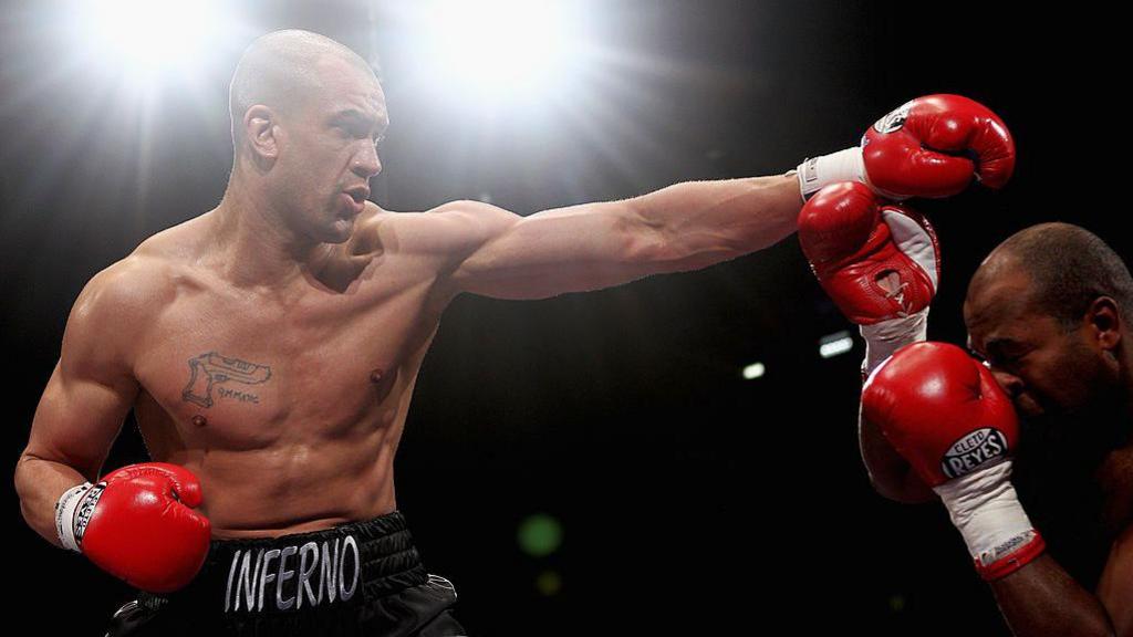 Two boxers face each other in a ring. The man on the left has his left arm stretched out in attack. He has a tattoo of a firearm on his chest with the word 9MMatic written next to it. The man on the right has his arms brought up in defence in front of his face as he ducks.