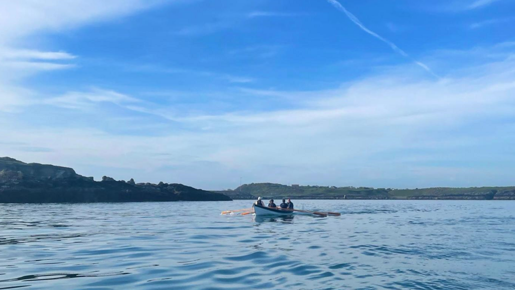 Group of people in a rowing boat on the blue water at Ardglass, County Down. A blue sky is above the coastline.
