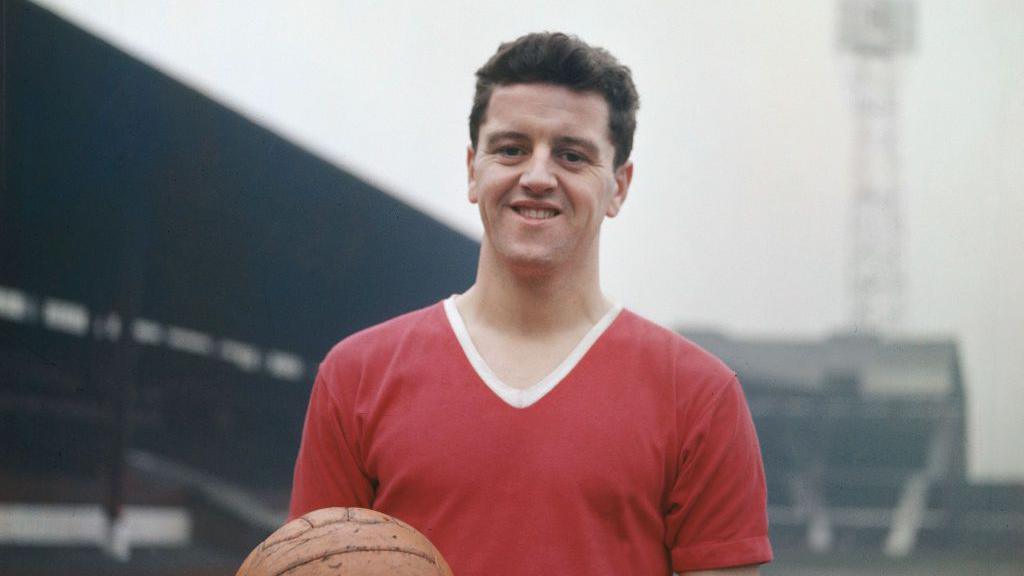 Footballer Tommy Taylor holding an orange football, dressed in a red and white top, standing inside a football ground