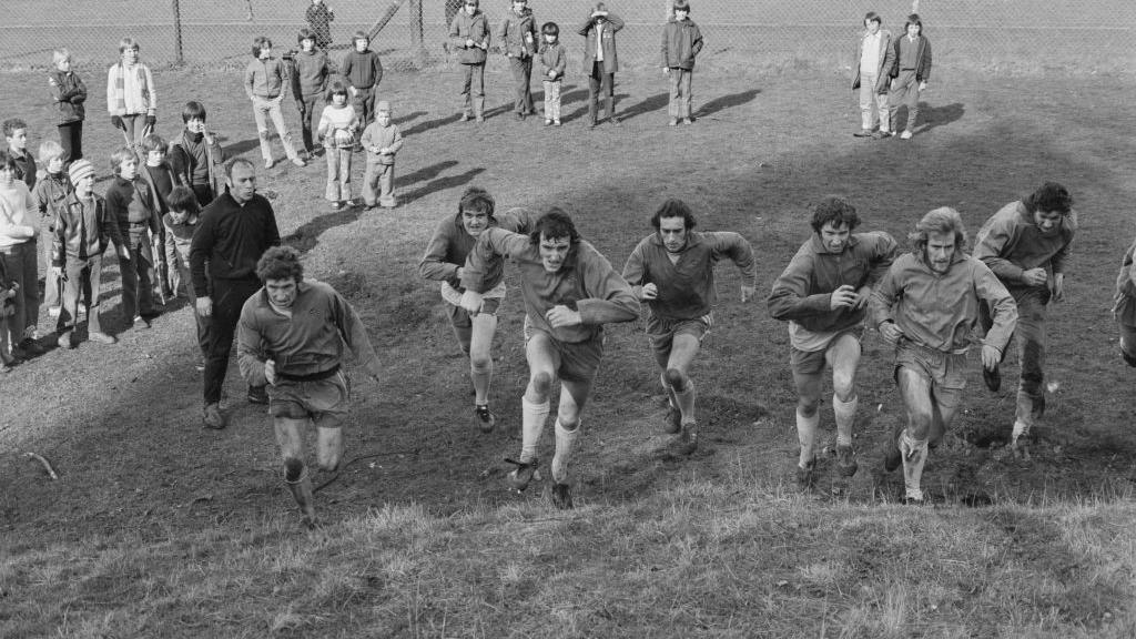 A black and white image of footballers running up a hill, towards the camera, as part of a training session. They are being watched by their coach, wearing a dark tracksuit, and about 20 children, in a picture taken in February 1973